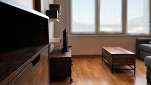 A modern living room with a wooden floor, a dark wooden desk, a grey sofa, and a window with blinds.