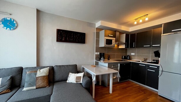 A modern kitchen with a dining table, grey sofa, and a clock on the wall.