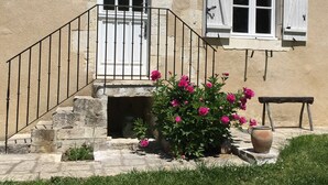 A stone building with a white door and windows, a metal staircase, and a flower bed with pink flowers. - Charming Riverside Cottage with Private Grounds, Fishing, Pet-Friendly, WiFi, and Parking (Mareuil-sur-Arnon)