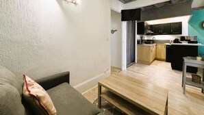 A compact living space with a sofa, a small wooden table, a kitchen area with black cabinets, and a visible door leading to another room. - Cozy 1BR Apartment in Mont-Dore Center, Near Thermes & Amenities, Sleeps 4 (Mont-Dore)
