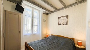 A bedroom with a bed, a wardrobe, a window with curtains, a wall-mounted TV, and a flower painting. - Cozy 1BR Apartment in Mont-Dore Center, Near Thermes & Amenities, Sleeps 4 (Mont-Dore)