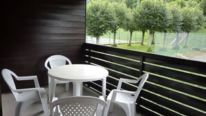A balcony with a round table and four white chairs, overlooking a grassy area with trees and a playground.