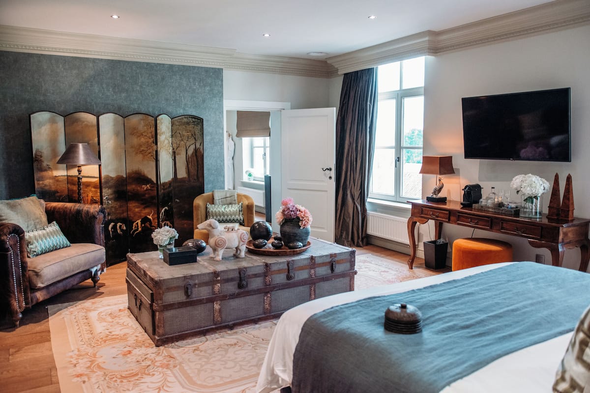 A spacious bedroom with a large bed, a vintage trunk as a coffee table, a leather sofa, and a desk with a TV.