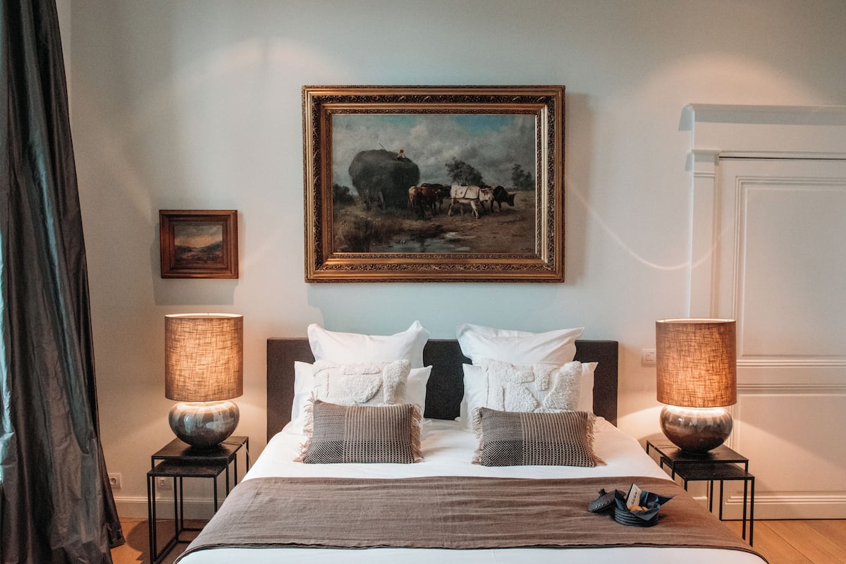 A hotel room with a large bed, two bedside lamps, a framed painting above the bed, and a small table with a lamp.