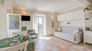Una sala de estar con un sofá, una mesa de comedor con sillas, un televisor montado en la pared y un ventanal corredizo que da a un área exterior.