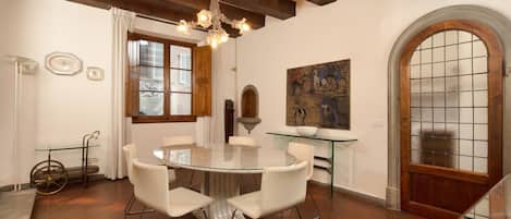A dining area with a round table, white chairs, a chandelier, and a painting on the wall.