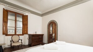 Un dormitorio con una cama grande, un aparador de madera, una silla y una ventana con contraventanas abiertas.
