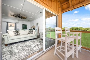 A room with a sliding glass door leading to a balcony, a white sofa with patterned cushions, a wooden ceiling, and a view of greenery.