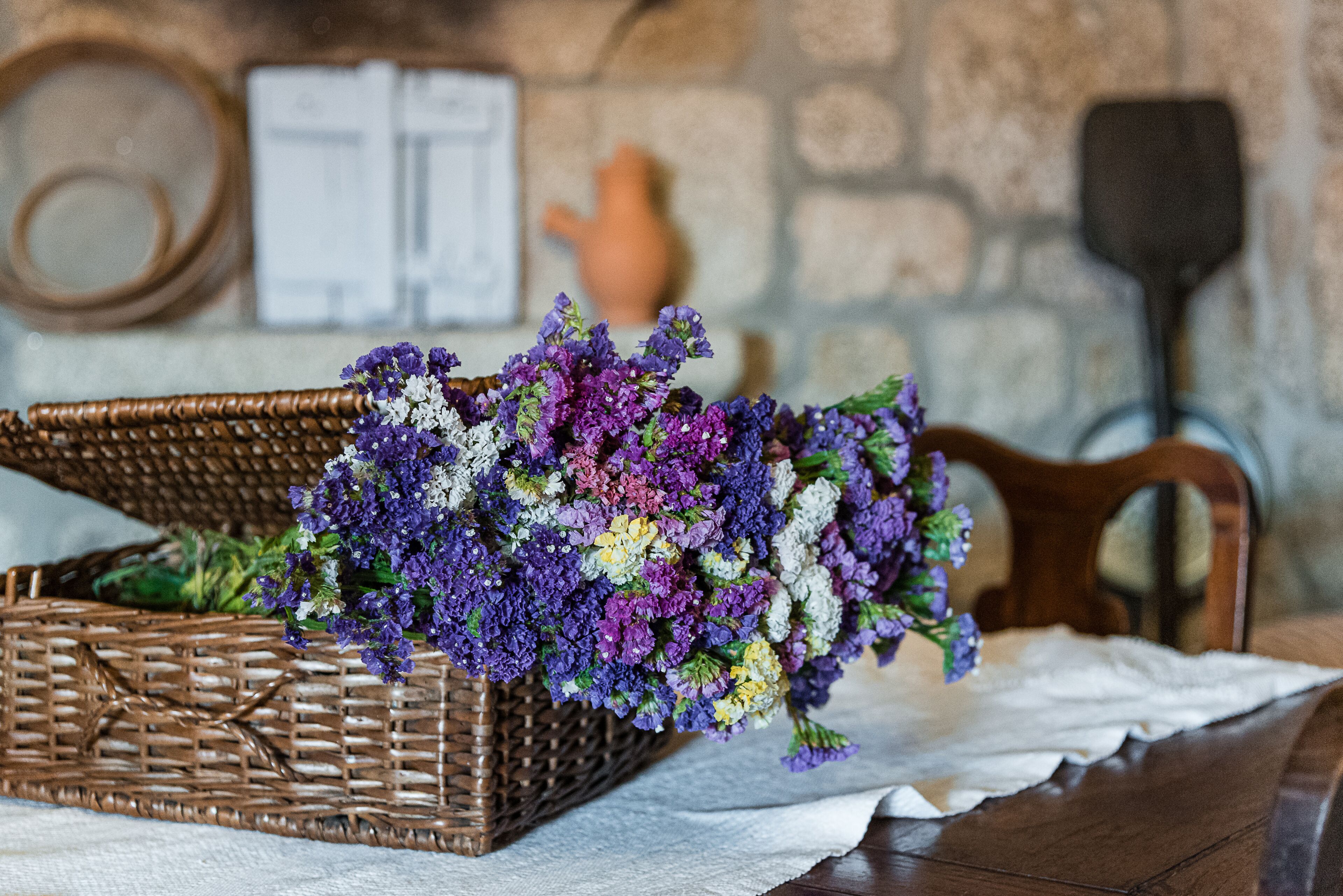 Un panier en osier rempli de fleurs violettes et blanches, posé sur une table en bois recouverte d’une nappe blanche.
