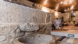 Una habitación de paredes de piedra con una gran pila de piedra, una mesa de madera con sillas y una araña.