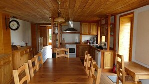 A wooden dining room with a long table and chairs, a kitchen area with wooden cabinets, and a clock on the wall. - Les hauts de Saint Christaud: large gite overlooking the Pyrenees (Saint-Christaud)