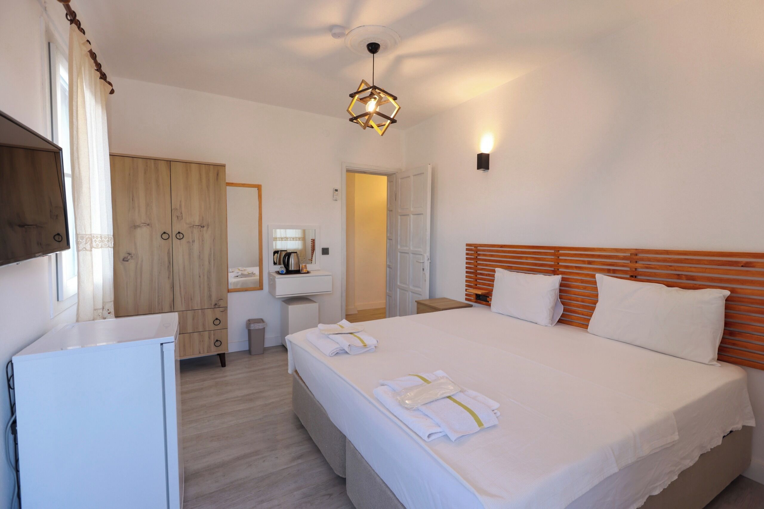Habitación de hotel con una cama grande, un armario de madera, un escritorio pequeño con hervidor y una lámpara colgante moderna.