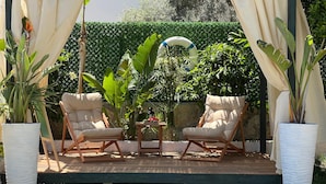 An outdoor seating area with two cushioned chairs and a small table, surrounded by lush greenery and a white curtain backdrop.