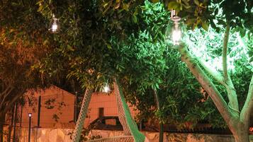 Un hamac est suspendu à un arbre dans une zone herbeuse, la nuit. On aperçoit un mur de pierre et un bâtiment en arrière-plan.