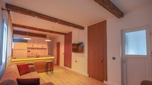 A modern living room with a wooden floor, a sofa, a dining area with a bar, and a kitchen in the background.