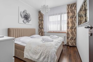 A bedroom with a bed, a bedside table, a window with curtains, a radiator, and a wall-mounted artwork.