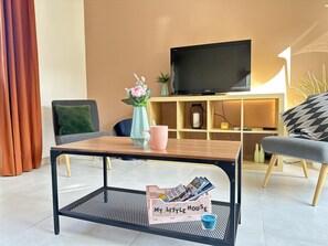 Ein modernes Wohnzimmer mit Couch, Fernseher auf einem Schrank, Couchtisch und einem kleinen Beistelltisch mit Vase und Tasse.