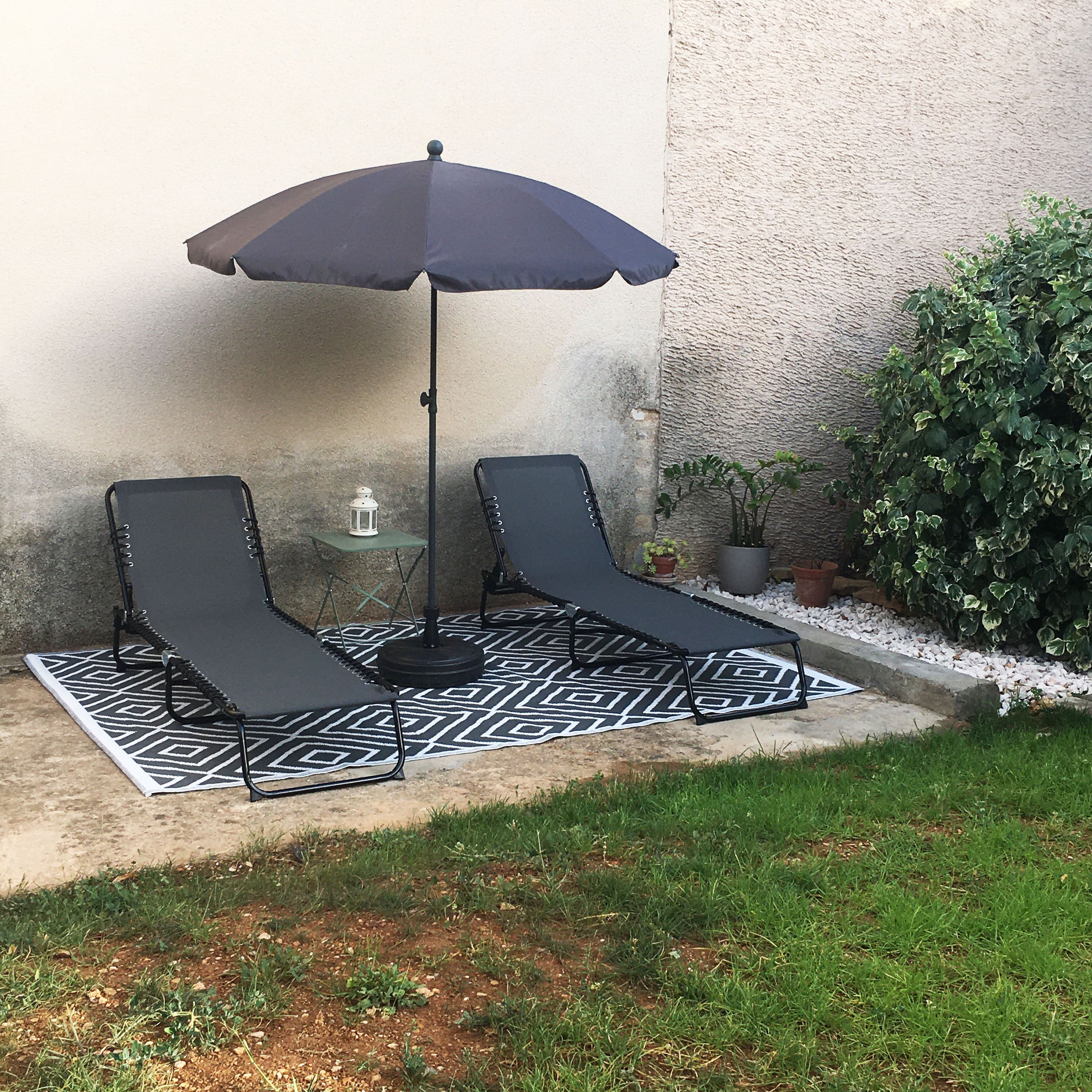 Twee lounge stoelen met een vloerkleed met patroon en een donkere parasol in een tuin.