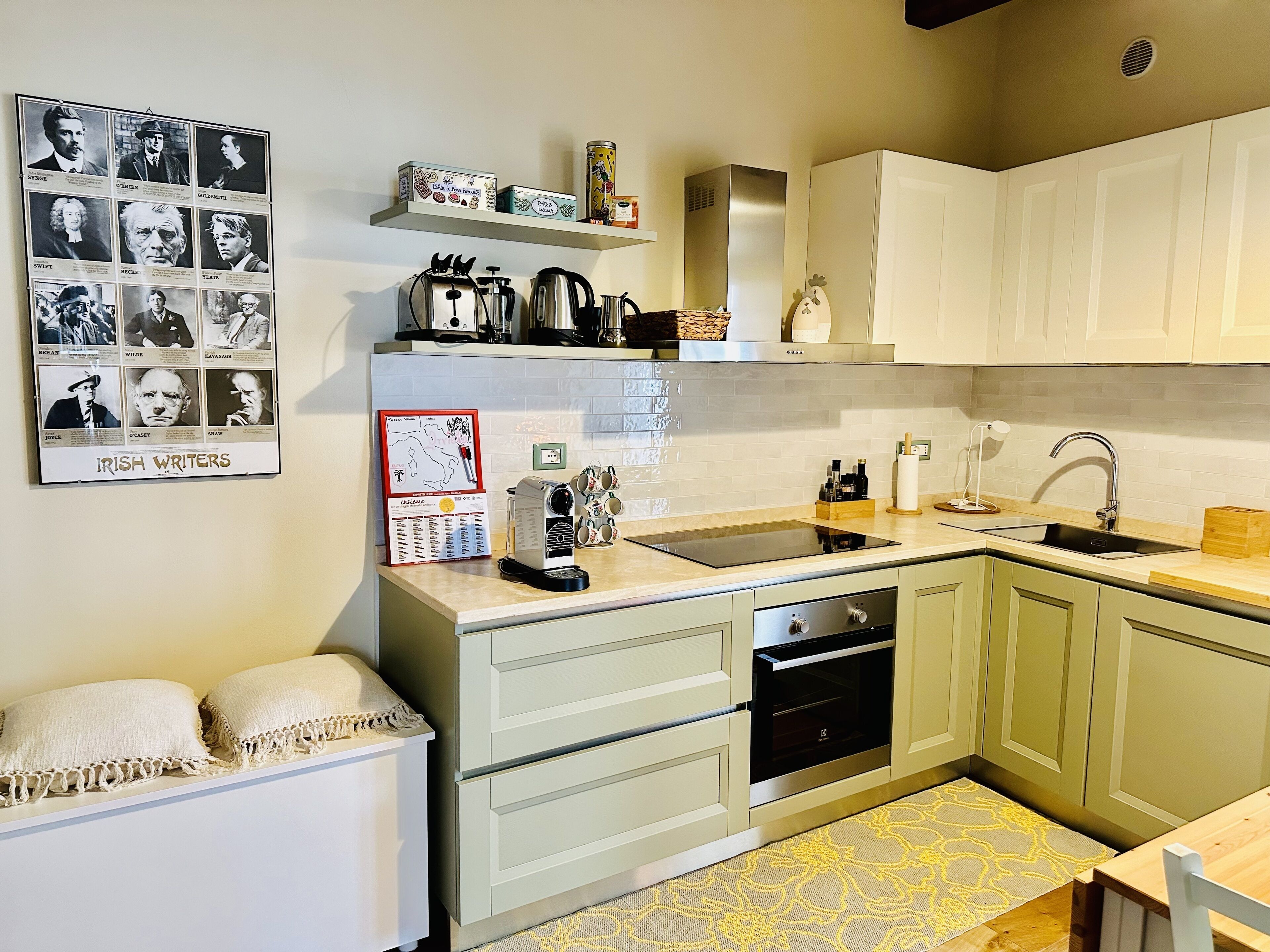 A modern kitchen with a built-in oven, a sink, and a coffee machine. There is a shelf with kitchen items and a framed poster on the wall.