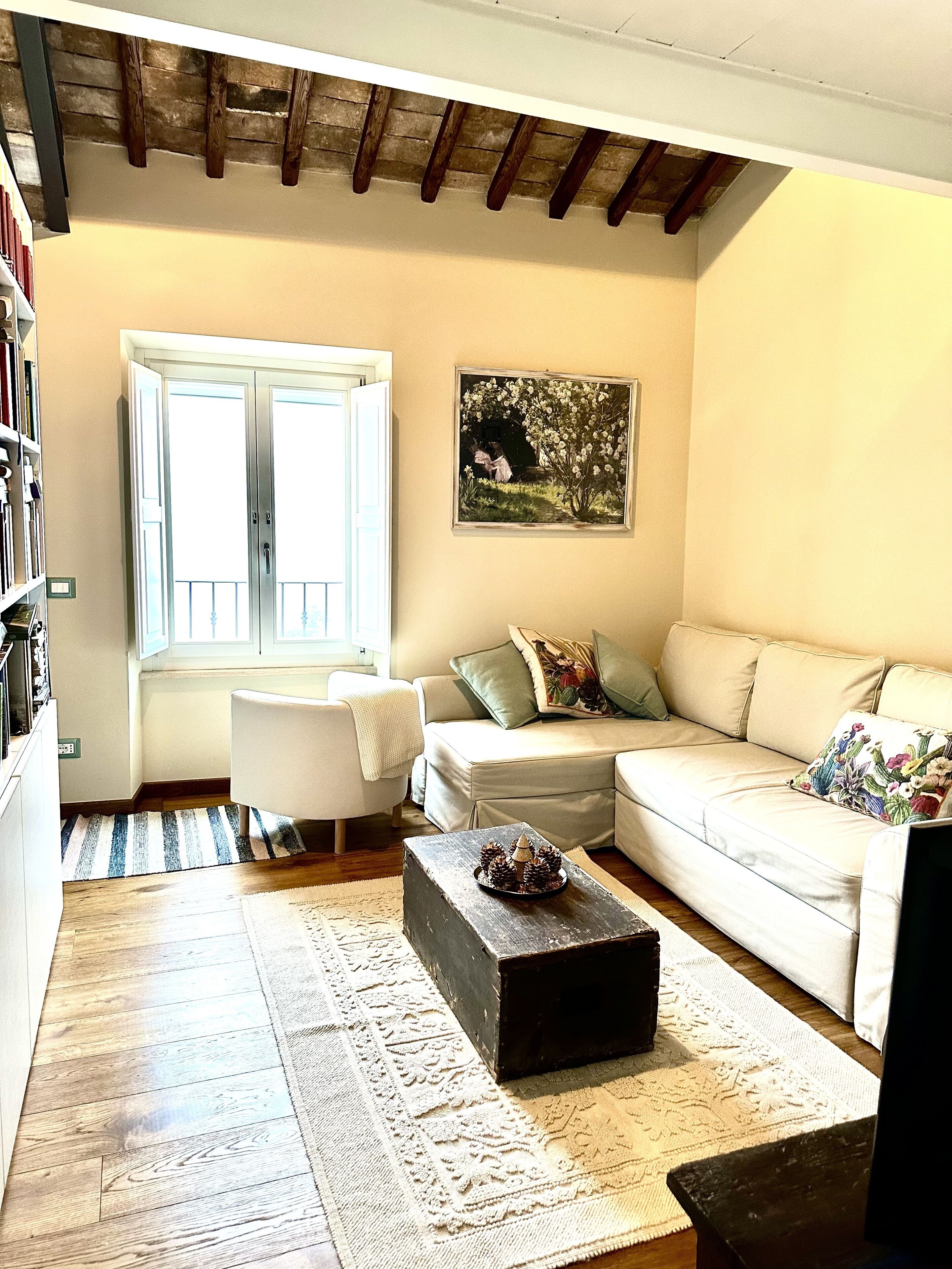 A living room with a sofa, a coffee table, a bookshelf, a window with blinds, and a painting on the wall.