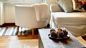 Un salon avec un canapé blanc, une table basse en bois ornée de pommes de pin, un tapis à rayures et un tableau représentant une scène de jardin.