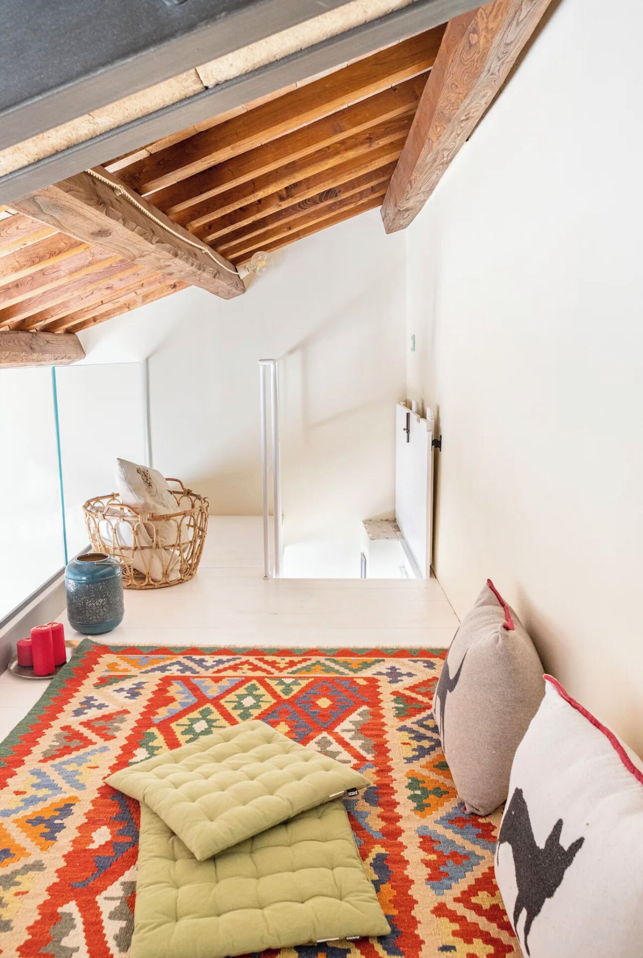 Una habitación con techo alto, vigas de madera a la vista, un tapete colorido con estampado y vistas a un balcón con cesta de mimbre y un florero azul.