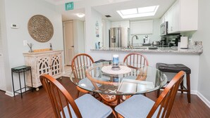Een eetgedeelte met een glazen tafel omringd door houten stoelen, een keuken met roestvrijstalen apparatuur, en een plafondventilator.