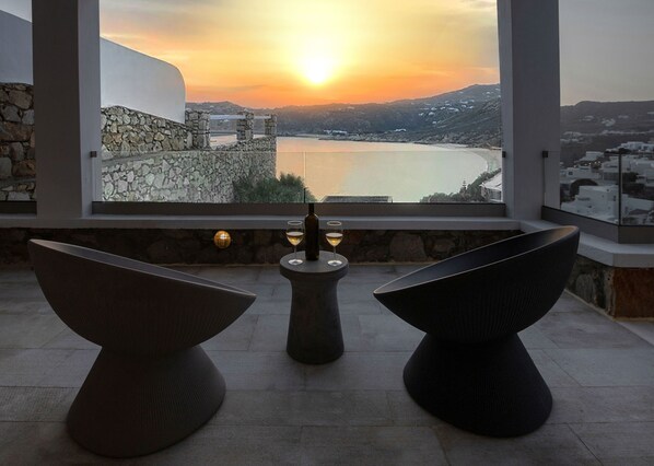 A terrace with two modern chairs, a table with a bottle and glass, and a view of a sunset over a body of water and hills.