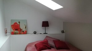 Een slaapkamer met dakraam, een bed met rode beddengoed, een lamp en een schilderij aan de muur.