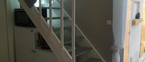 Un escalier menant à un palier où se trouvent un four à micro-ondes et des livres sur une étagère, une table en bois et un tapis avec une flèche rouge sur le sol.