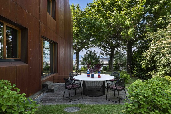 Un espacio moderno al aire libre con una mesa redonda y sillas, rodeado de exuberante vegetación y vistas a un cuerpo de agua.