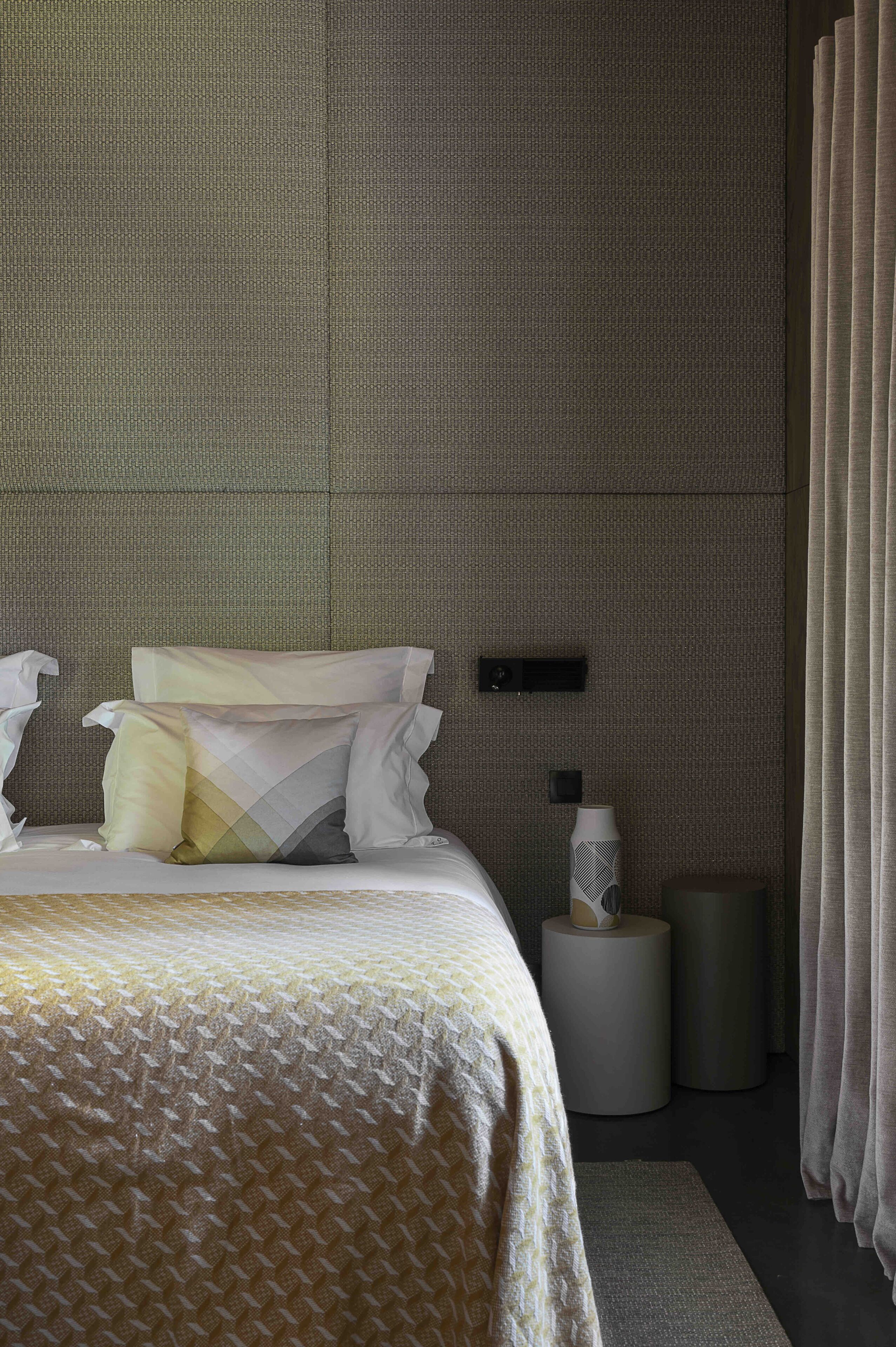 A modern hotel room with a large bed, a bedside table, a lamp, and a wall-mounted control panel.