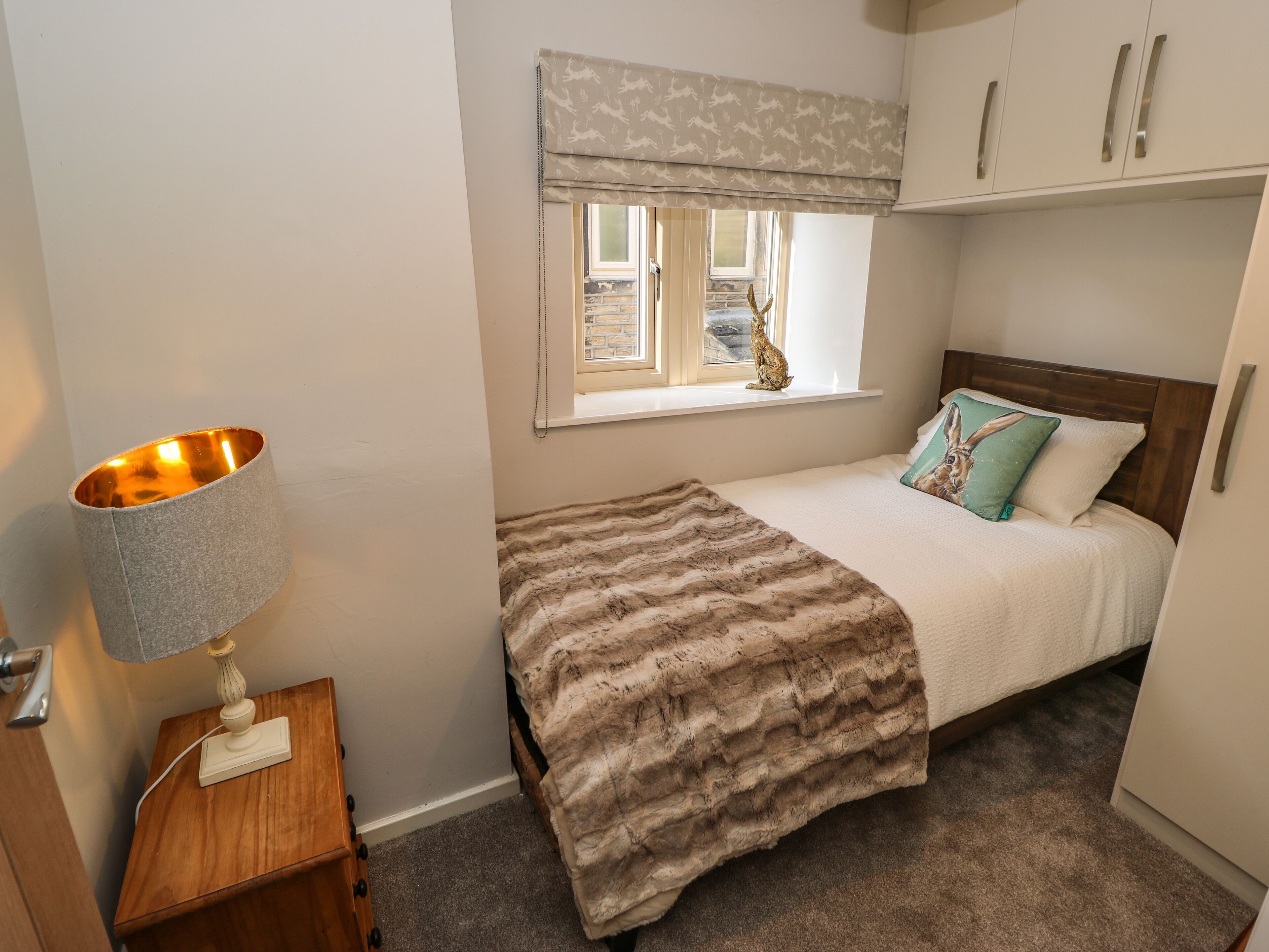 A bedroom with a bed, a nightstand with a lamp, a window with a view, and a shelf with decorative items.