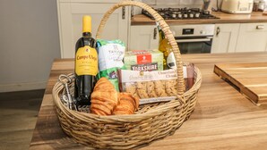 A wicker basket on a kitchen counter containing a bottle of Campo Viejo Rioja wine, a pack of Taylor's of Harrogate tea, a box of Grandma Wild's chocolate chunk biscuits, and croissants. - Kingfisher Cottage (Holmfirth)