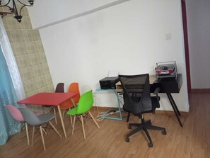 A room with a dining table and chairs, a black office chair, a desk with a printer, and a small shelf with books.