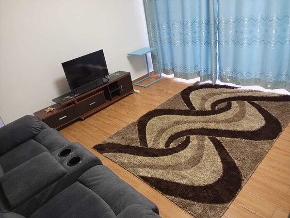 Una sala de estar con un sofá, un televisor sobre un mueble de madera, una mesa azul y una alfombra estampada.