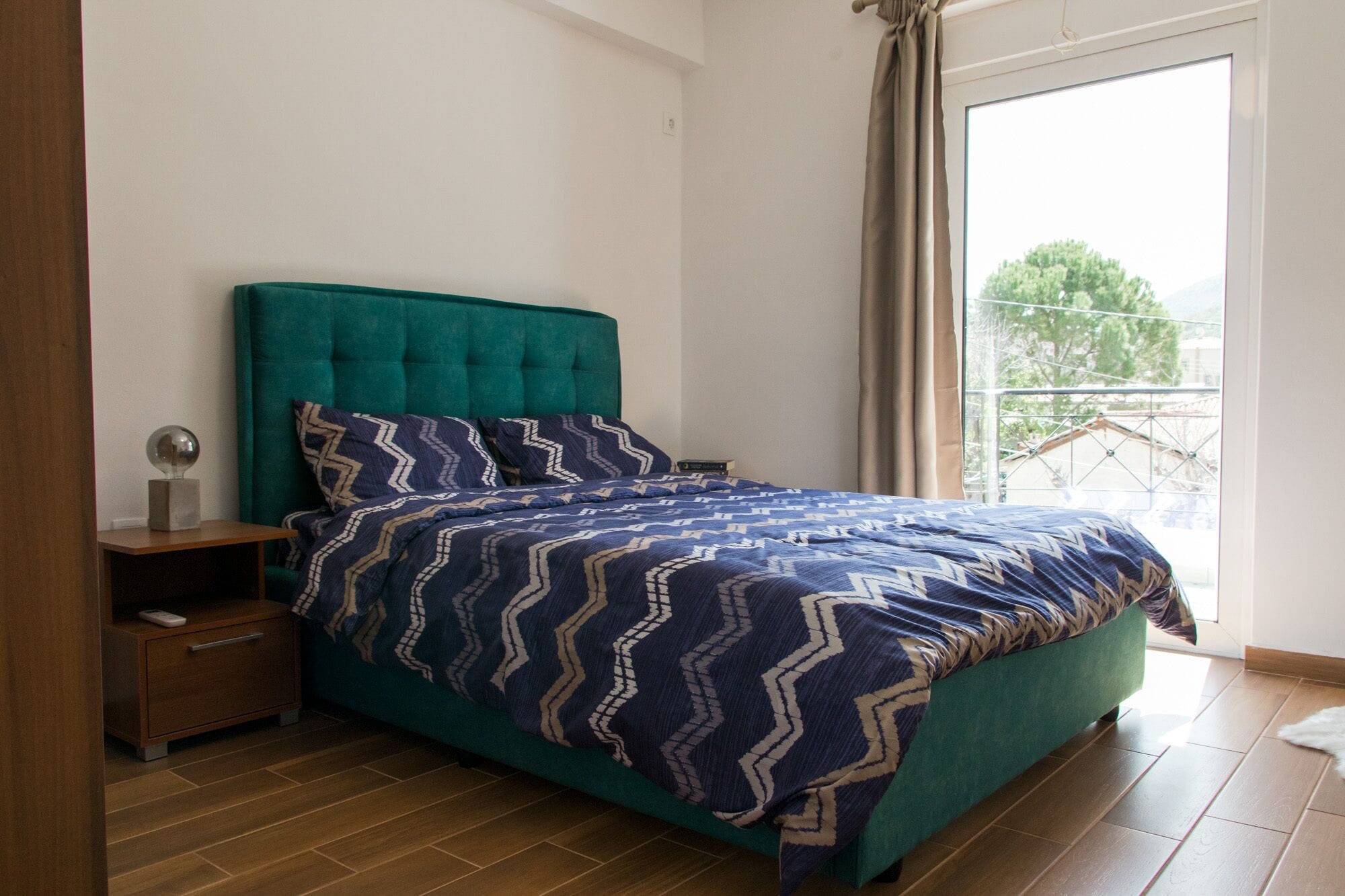 A bedroom with a teal upholstered headboard, a patterned bedspread, a bedside table with a lamp, and a view of a balcony with a tree.