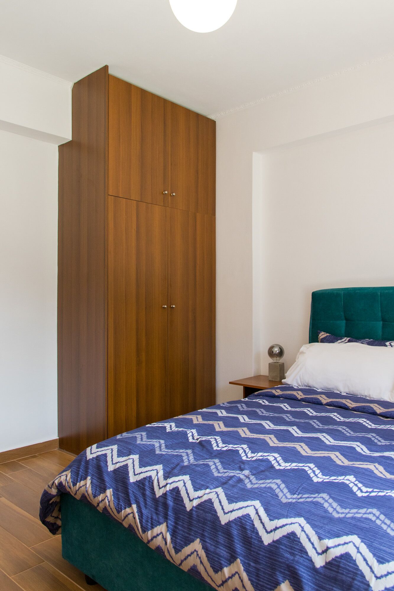 Une chambre avec une armoire en bois, un lit recouvert d’une couvre-lit à motifs et une tête de lit recouverte d’un tissu turquoise.