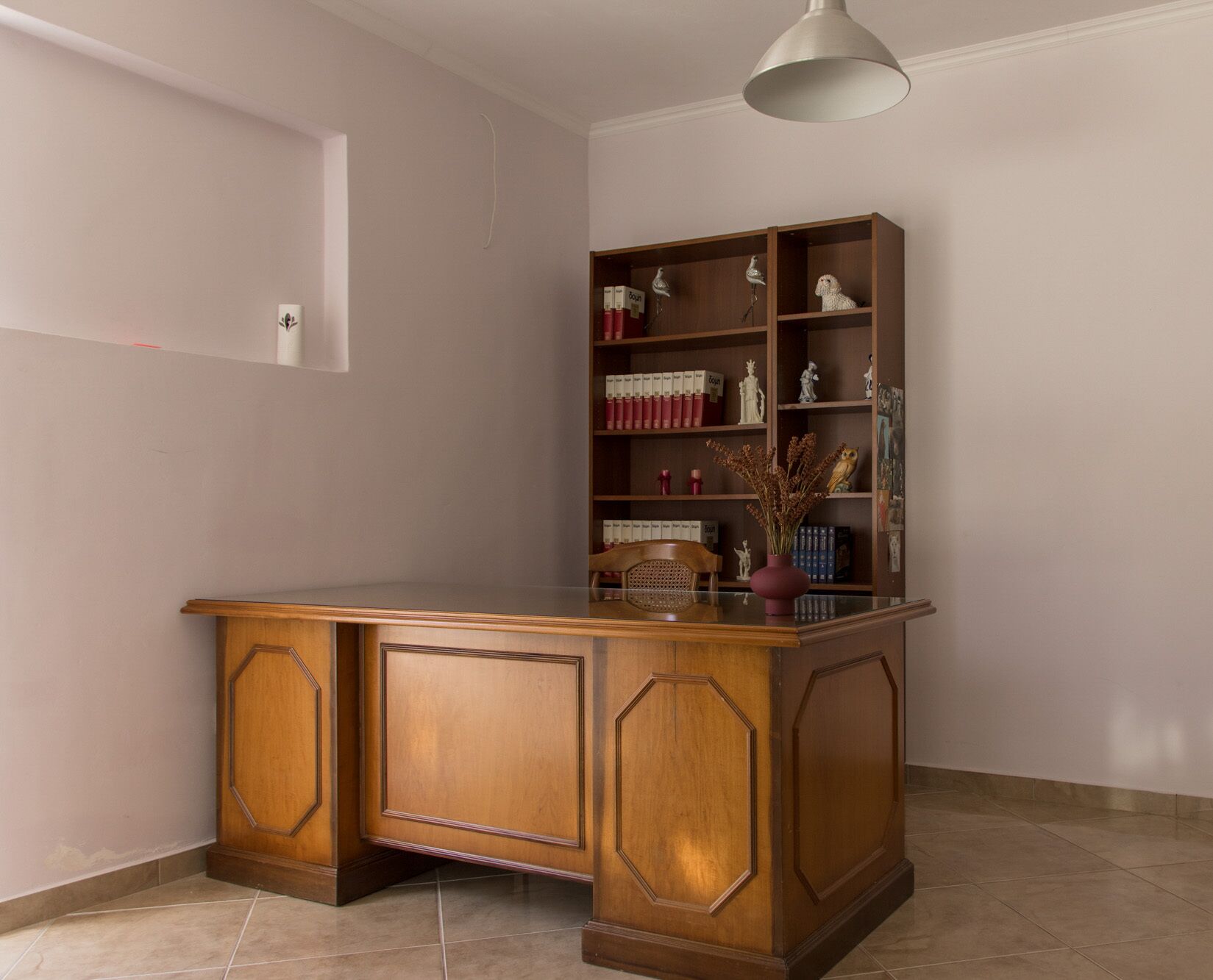 A wooden desk with a glass top, a bookshelf with decorative items, and a hanging light fixture.