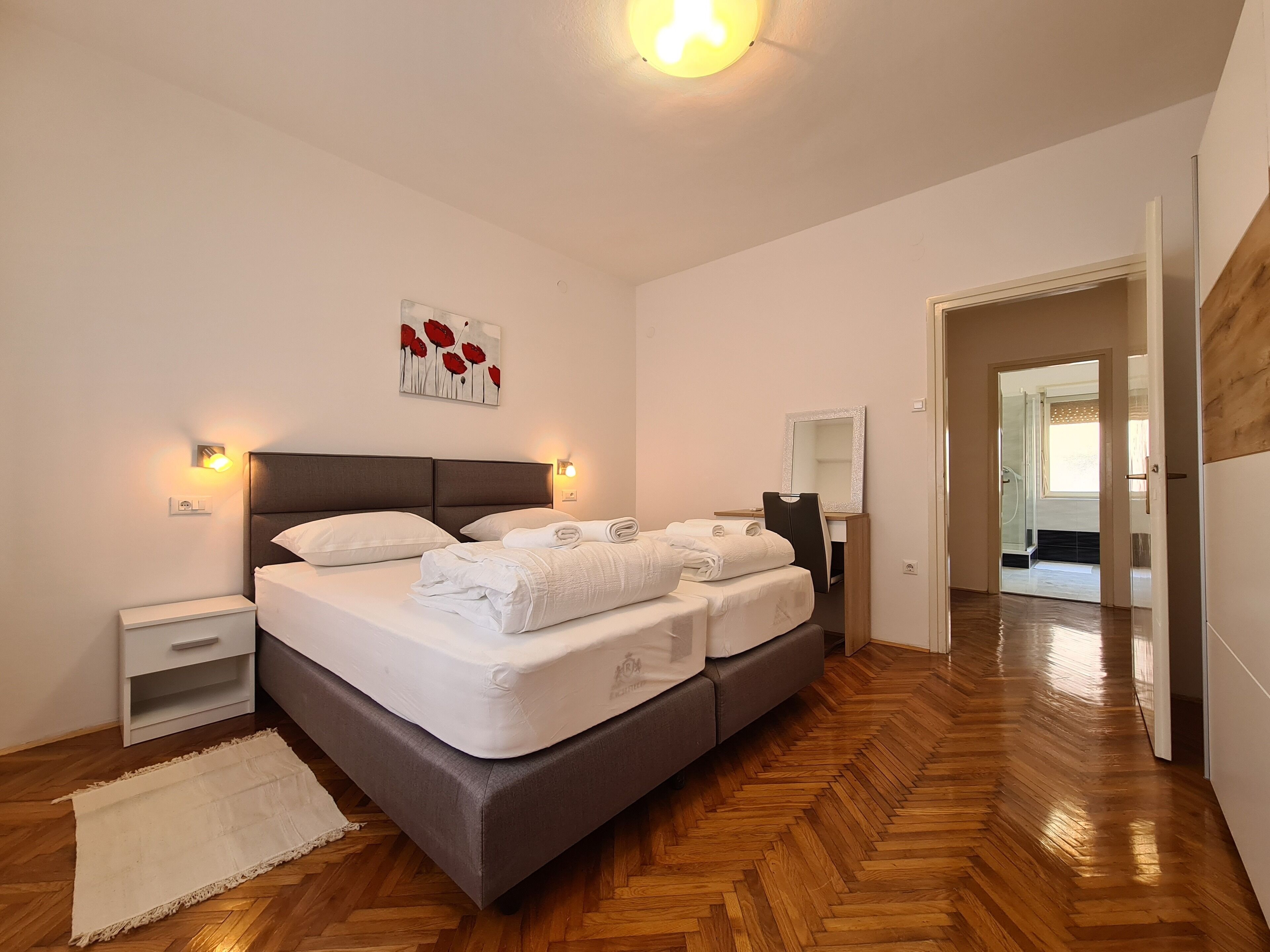 Een slaapkamer met een groot bed, nachtkastjes, een bureau met stoel, een spiegel en een deur naar een andere kamer.