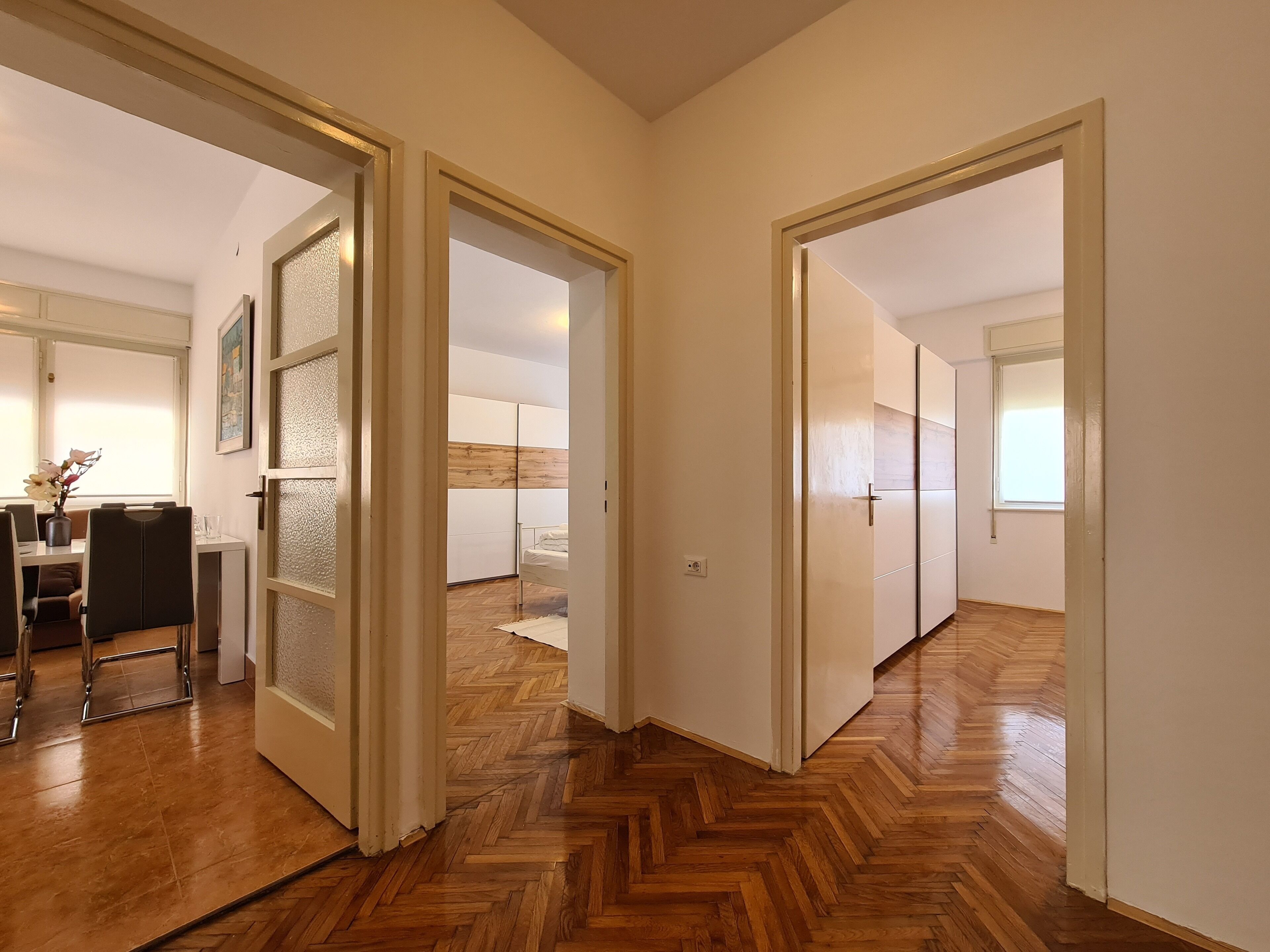 Un couloir au sol en bois et aux murs blancs, qui mène à une chambre à coucher équipée d’un lit et d’une table de chevet.
