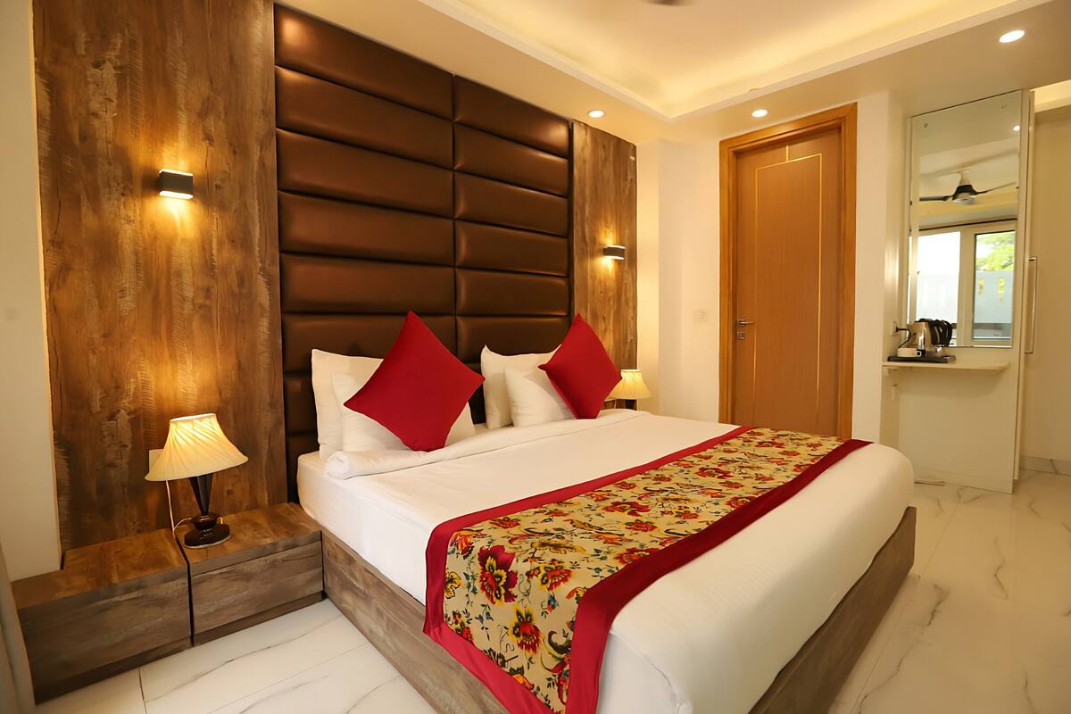 Un dormitorio moderno con una cama grande, cabecera de madera, almohadas rojas y blancas, colcha con estampado floral, mesita de noche con lámpara y espejo sobre la puerta.