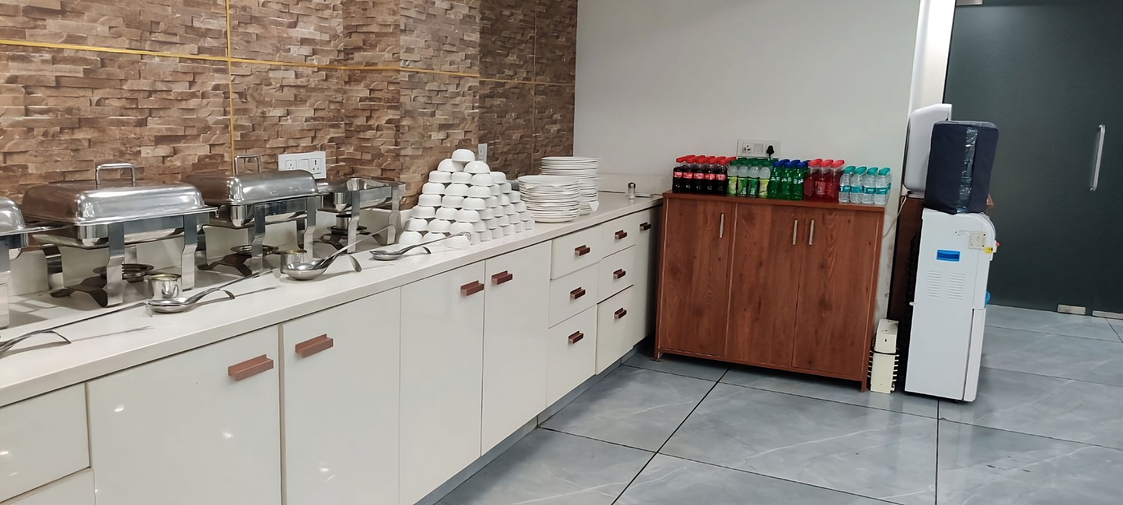 A buffet setup with stainless steel chafing dishes, a stack of white plates, and a cabinet with a water dispenser.