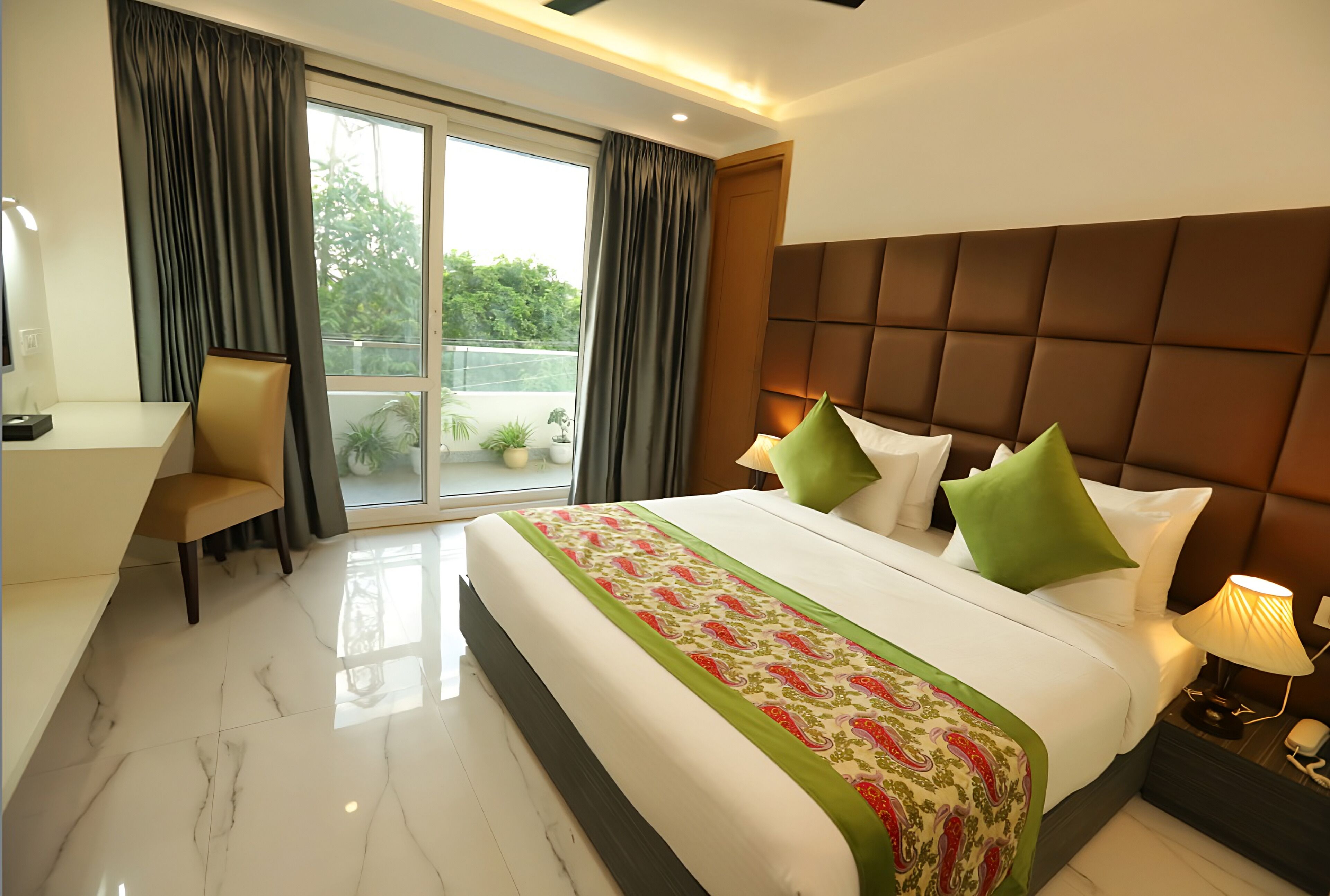 A hotel room with a large bed, a desk, and a chair. There is a window with curtains, a view of greenery outside, and a padded headboard.