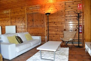 A room with wooden walls, a white sofa with cushions, a small table, a chair, a floor lamp, and a fire extinguisher.