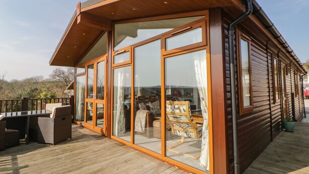 A wooden cabin with large glass doors and windows, a deck with outdoor furniture, and a view of the surrounding landscape. - Meadow Lodge (Plymouth)
