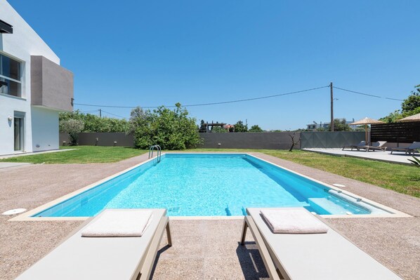 A rectangular swimming pool with a clear blue surface, surrounded by a paved deck. There are two lounge chairs with towels on them.