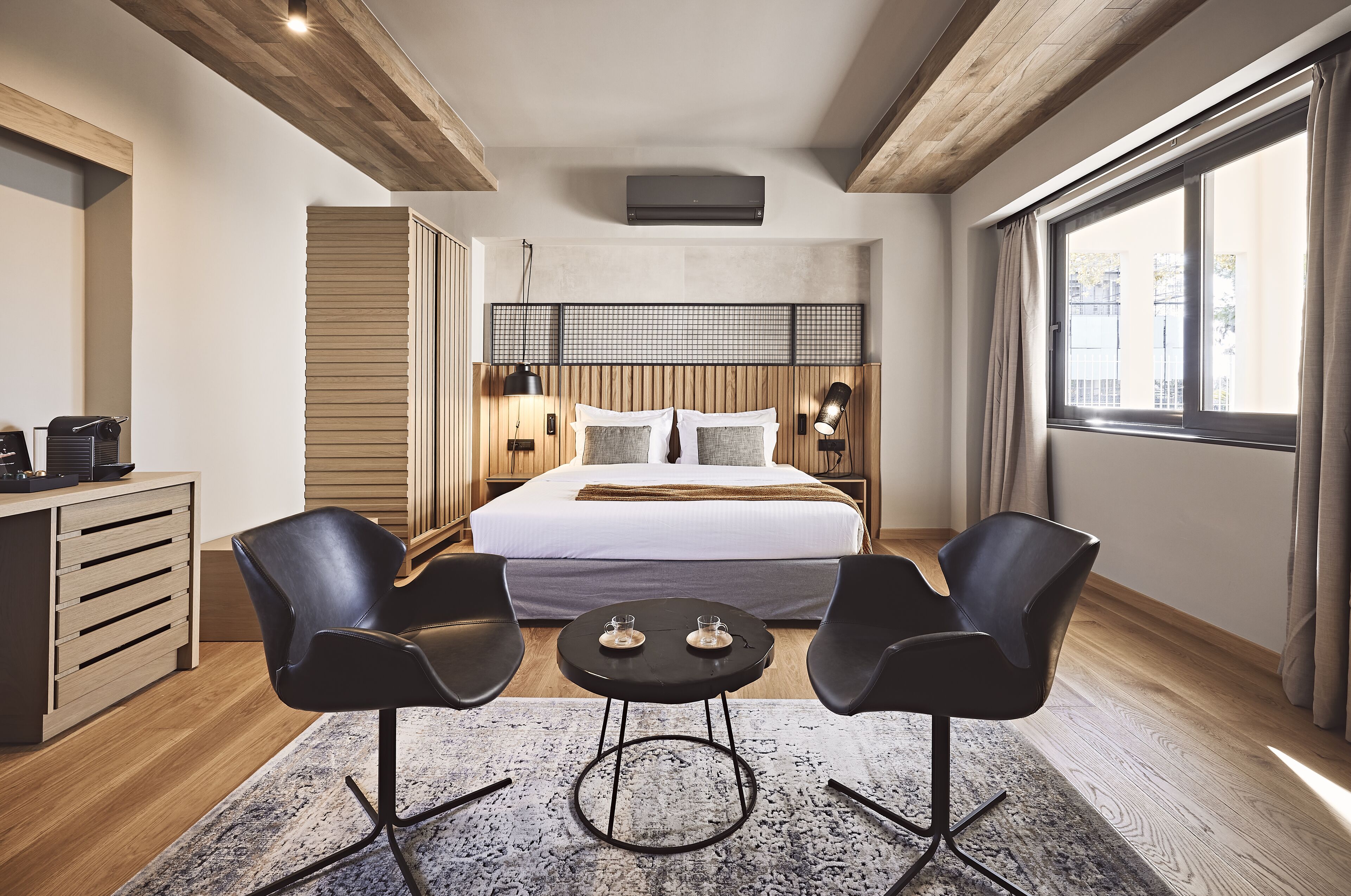 A modern hotel room with a large bed, two black chairs, a small table, a wooden desk, and a large window with curtains.