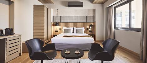 A modern hotel room with a large bed, two black chairs, a small table, a wooden desk, and a large window with curtains.