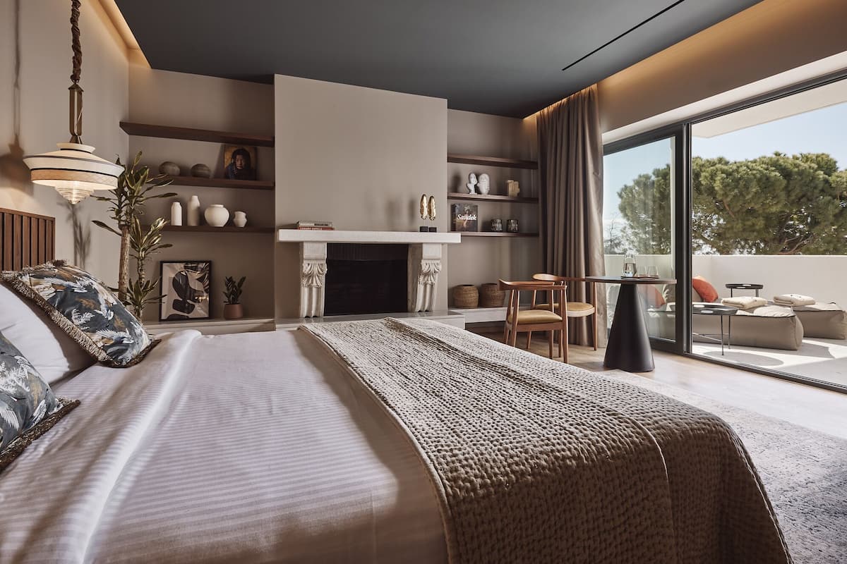 A modern bedroom with a large bed, a fireplace, a dining area, and a balcony with a view of trees.